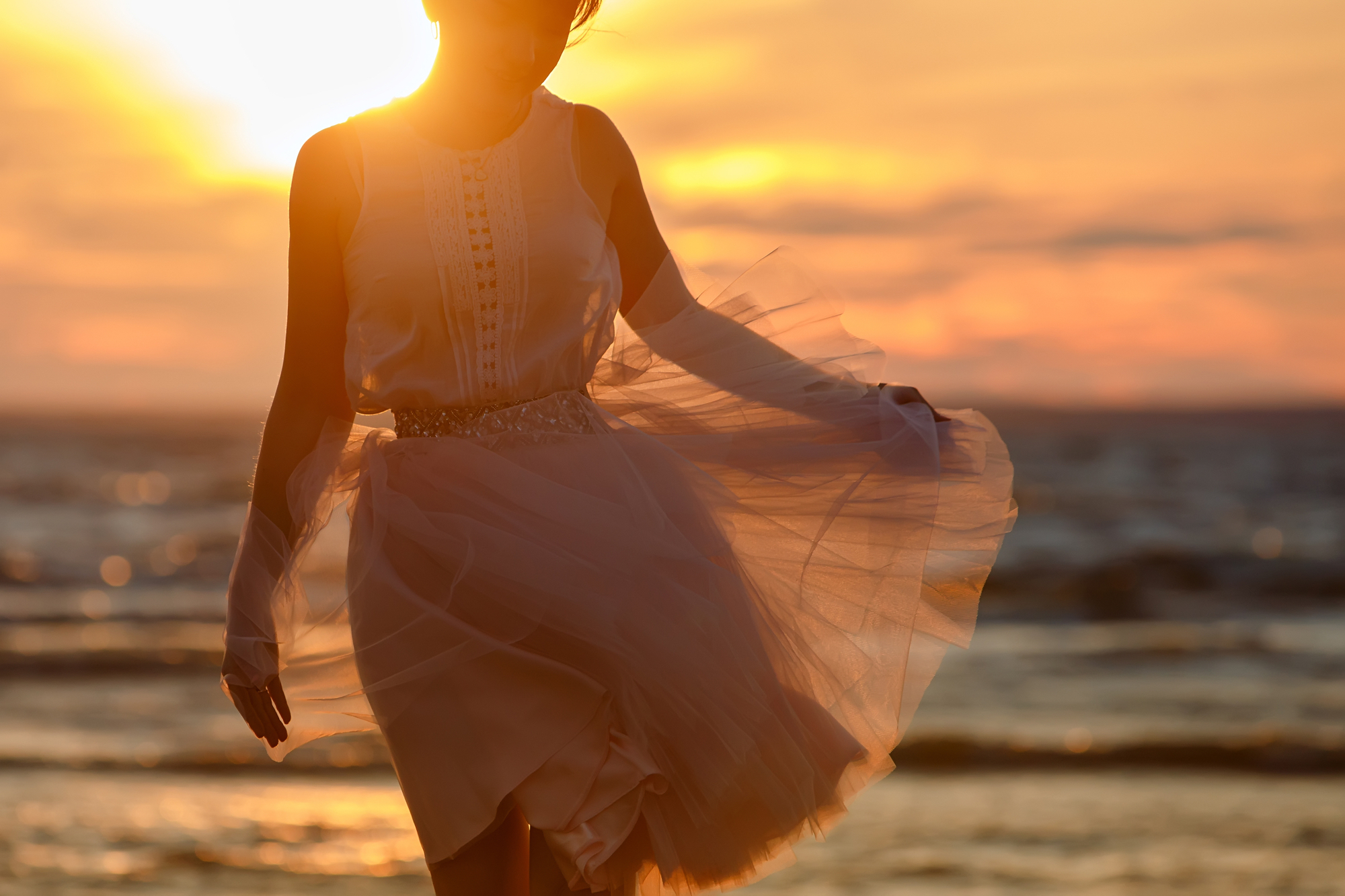 The silhouette of a slim girl in lush pink short skirt, standing on the sand against the sea and the Golden sunset